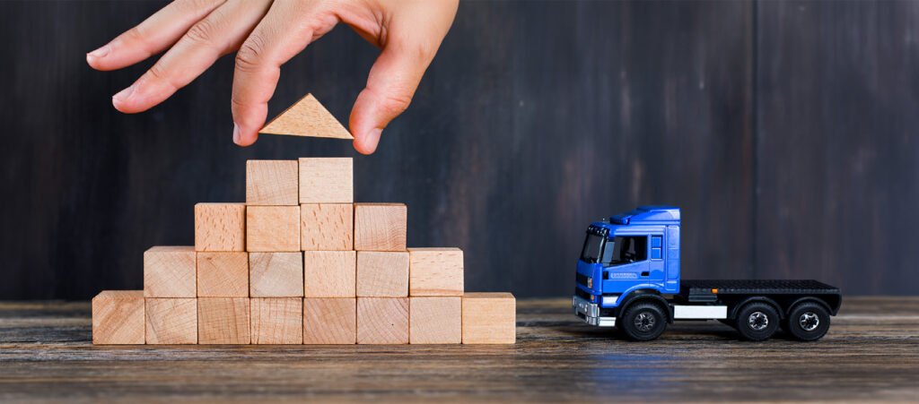 Een hand plaatst het bovenste blokje op een trap van houten blokken, met rechts een blauwe speelgoedvrachtwagen